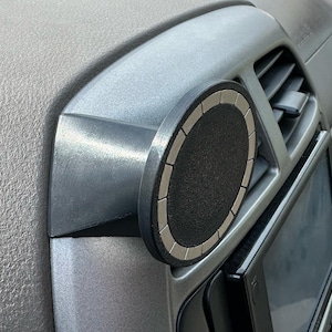 May include: A silver car vent mount with a black and silver circular phone holder. The mount is attached to the dashboard vent, with a black screen visible below. The holder has a textured surface.