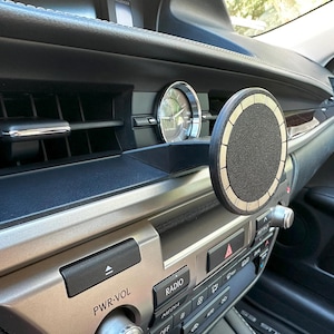 Op de afbeelding: Zwarte en gouden telefoonhouder voor in de auto bevestigd aan het dashboard van een auto. De houder heeft een cirkelvormig ontwerp met een getextureerd midden en een goudkleurige rand. Het dashboard heeft een klok, ventilatieopeningen en radioknoppen.