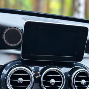 Puede incluir: Primer plano del salpicadero de un coche con una pantalla rectangular, rejillas de ventilación y un altavoz circular. El salpicadero es negro y plateado, destacando la tecnología y el diseño interior del vehículo.