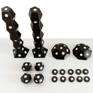 May include: A collection of black geometric components featuring white circular accents. The set includes two tall stacked towers, two dome-shaped pieces, four small multifaceted polyhedrons, and ten flat circular discs arranged in two rows.