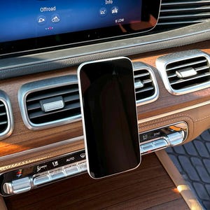 Puede incluir: Un teléfono inteligente negro está montado en el salpicadero de un coche. El salpicadero presenta molduras de veta de madera, rejillas de ventilación y botones de control. La pantalla de infoentretenimiento muestra información. El interior del coche tiene una combinación de colores marrón y negro.