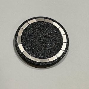 May include: A round, black coaster with a textured surface and a silver-colored border. The coaster has a series of rectangular silver accents around the outer edge. The coaster is designed to protect surfaces from drink rings.