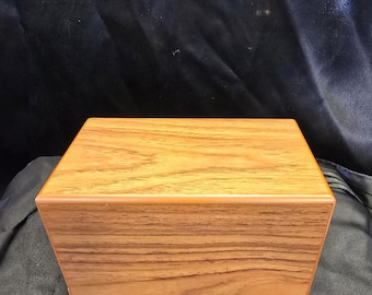 Peut inclure: Une boîte rectangulaire en bois avec une finition brun clair et un grain de bois visible. La boîte a un design simple et épuré avec un dessus et des côtés plats. La boîte est posée sur un tissu bleu foncé.