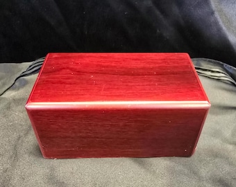 Peut inclure: Une boîte rectangulaire en bois avec une finition brun-rougeâtre. La boîte présente un design simple et épuré avec une surface lisse et polie. Les bords sont légèrement arrondis, et la boîte semble bien fabriquée.