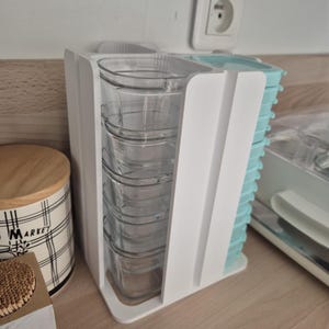 Peut inclure: Un organisateur de conteneurs en plastique blanc contenant des récipients de stockage alimentaires en verre transparent. L'organisateur a une section pour les conteneurs et une autre pour les conteneurs à couvercles bleus. Un récipient décoratif avec un couvercle en bois est à gauche.