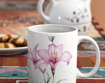Könnte beinhalten: Weiße Keramiktasse mit rosa Blumendesign, eine weiße Teekanne und ein Teller mit Keksen. Die Tasse zeigt eine Aquarellillustration von rosa Lilien. Der Hintergrund umfasst eine farbenfrohe gemusterte Tischdecke. Der Text "Ships Quick!" ist im Bild.