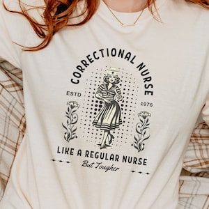 May include: A beige t-shirt with a black and white graphic design. The graphic features a woman in a nurse's uniform with the text "Correctional Nurse Est'd 1976 Like a Regular Nurse But Tougher".