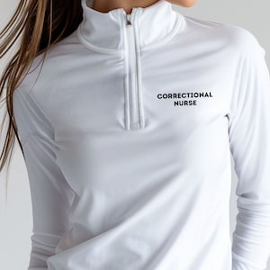 May include: A white long-sleeve shirt with a quarter-zip collar. The text "CORRECTIONAL NURSE" is printed in black on the upper chest. The shirt is made of a smooth, lightweight fabric.