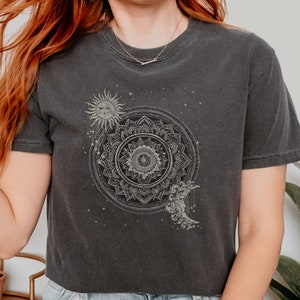 May include: A black cropped t-shirt with a white mandala design featuring a sun, moon, and stars. The mandala is surrounded by a circle of stars.