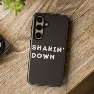 Könnte beinhalten: Eine schwarze Smartphone-Hülle mit dem weißen Schriftzug "SHAKIN' DOWN". Die Hülle hat eine Dreifach-Kamera und liegt auf einer Holzoberfläche. Die Hülle dient zum Schutz des Telefons.