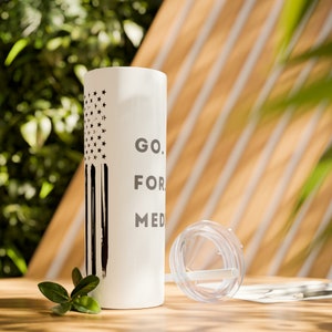 May include: White tumbler with a black and white American flag design and the text "GO. FOR. MED." printed on the side. The tumbler is sitting on a wooden surface with a clear lid and straw next to it.