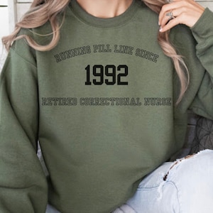 May include: Green sweatshirt with the text "Running Pill Line Since 1992 Retired Correctional Nurse" printed on the front.