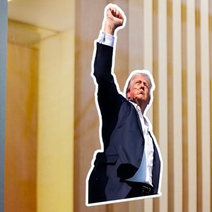 May include: A man in a dark blue suit with a white shirt raises his fist in the air. He is standing in front of a building with vertical lines.