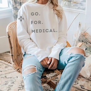 May include: A white sweatshirt with the text "GO. FOR. MEDICAL." in gray block letters. A small logo is on the left sleeve. The person is wearing light blue jeans with distressed knees and is seated in a wicker chair.