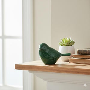 May include: A dark green ceramic bird ornament. The bird is positioned on a wooden surface, with a succulent in a white pot and books in the background. The bird has detailed wing and tail feathers.