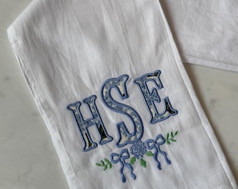 May include: White linen hand towel embroidered with the letters "HSE" in blue floral print. The design includes blue bows, green leaves, and a cluster of blue flowers. The towel is folded.