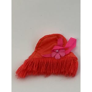 Puede incluir: Un pequeño sombrero naranja con un borde con flecos y un acento de flor y cinta rosa. El sombrero está hecho de una tela suave y tiene una forma redondeada. La flor rosa tiene una perla en el centro.