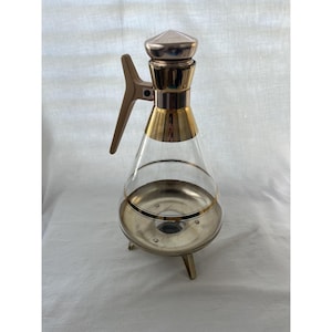 May include: A vintage glass and metal decanter with a conical shape. The decanter features a gold-colored band and accents, a silver-colored base, and a matching lid. It has a brown handle and stands on three small legs.