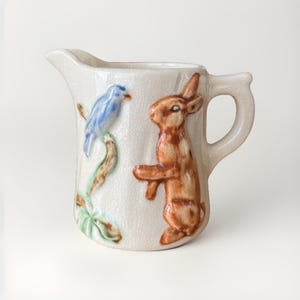 Puede incluir: Jarra de cerámica color crema con diseño de pájaro azul y conejo marrón. La jarra tiene asa y pico vertedor. El diseño está en relieve.