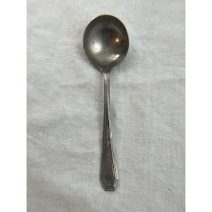 May include: A silver spoon with a round bowl and a long, decorative handle. The spoon is made of a shiny, metallic material and rests on a white, textured surface. The handle has a geometric design near the end.