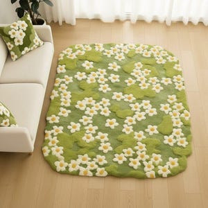 Könnte beinhalten: Ein grüner Teppich mit einem Blumenmuster aus weißen und gelben Blüten. Der Teppich hat ein weiches, strukturiertes Aussehen und liegt auf einem Holzboden. Ein passendes Kissen liegt auf einem weißen Sofa.