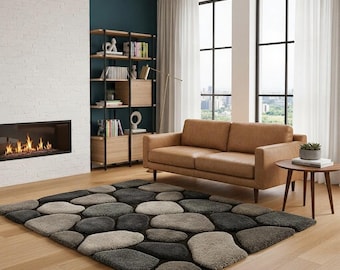May include: A stone-effect rug in a living room. The rug features a pattern of grey, black, and beige stones. A brown leather sofa and a small round table are also visible. The rug is placed on a wooden floor.