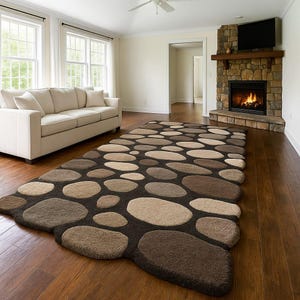 Puede incluir: Una alfombra rectangular grande con un diseño de guijarros en tonos marrones, beige y beige claro. La alfombra está colocada sobre un suelo de madera en un salón, con un sofá y una chimenea en el fondo.