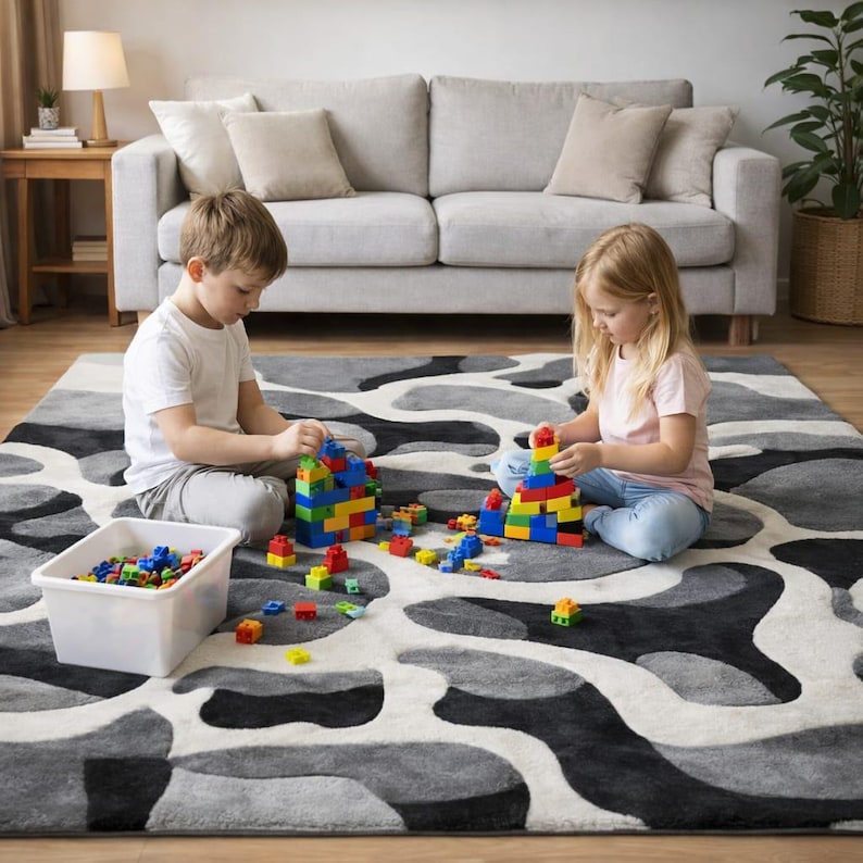 May include: Two children are playing with colorful building blocks on a patterned rug. The rug has a gray, black, and white abstract design. A white plastic bin holds additional blocks. A gray sofa is in the background.