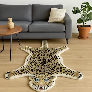 Puede incluir: Alfombra con estampado de leopardo beige y marrón con un diseño de rostro detallado. La alfombra tiene forma de piel de leopardo, con un fondo color crema y manchas negras y marrones. La cara tiene ojos azules y nariz rosa.