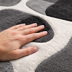 May include: A close-up of a soft, patterned rug with a hand resting on it. The rug features a design of overlapping shapes in shades of gray and white. The texture appears plush and comfortable.