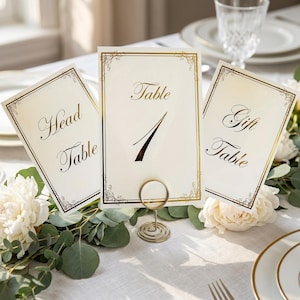Puede incluir: Elegantes tarjetas de números de mesa con bordes dorados y caligrafía. Las tarjetas dicen "Head Table", "Table 1" y "Gift Table". Un soporte de alambre dorado sostiene la tarjeta "Table 1". Flores blancas y vegetación decoran la mesa.