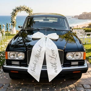 Könnte beinhalten: Ein schwarzes Oldtimer-Auto, geschmückt mit einer großen weißen Schleife und Bändern mit den Aufschriften "Sarah & Michael" und "with Love". Das Auto steht in der Nähe einer Strandhochzeit.