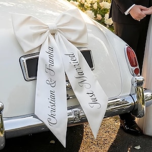 Könnte beinhalten: Eine große, cremefarbene Satinschleife an der Rückseite eines weißen Oldtimers. Die beiden langen, herabhängenden Bänder sind mit schwarzem Text bedruckt. Auf dem linken Band steht Christian & Franka, auf dem rechten Just Married in Schreibschrift.