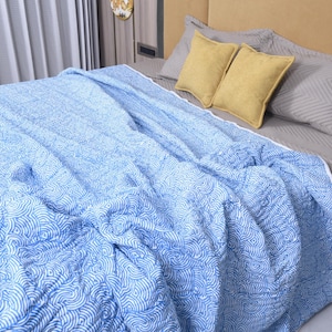 May include: A light blue and white quilted bedspread featuring a repeating wave pattern. The fabric has a soft, textured appearance with a white border edge, draped across a bed with grey bedding and two mustard yellow square throw pillows.