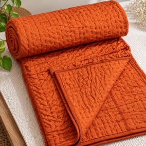 May include: A burnt orange quilted blanket featuring a textured, crinkled surface with visible grid-like stitching throughout. The blanket is partially rolled at the top, revealing a folded corner with a finished, solid-colored fabric edge.