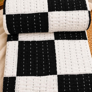 May include: A folded quilt featuring a high-contrast black and white checkerboard pattern. The fabric is detailed with visible white running stitch quilting lines that create a textured, grid-like appearance across the entire surface.