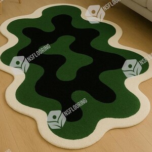 May include: A modern, abstract area rug with a unique, organic shape. The rug features a pattern of dark green, medium green, and black shapes, outlined with a cream-colored border. The rug is placed on a light-colored wooden floor.