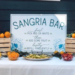 Puede incluir: Un cartel blanco con el texto "SANGRIA BAR" en letras verdes. El cartel también incluye instrucciones para hacer sangría: "first... PICK RED OR WHITE... then... ADD SOME FRUIT... lastly... REFILL FREQUENTLY". El cartel se muestra en una mesa con dos dispensadores de bebidas de vidrio, uno lleno de vino tinto y el otro de vino blanco. También hay tazones de fruta, incluyendo limas, limones, naranjas, fresas y arándanos.