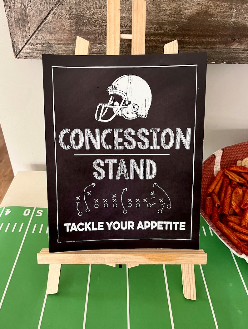 Puede incluir: Un letrero negro de pizarra con texto blanco que dice "Concession Stand" y "Tackle Your Appetite". Un gr&aacute;fico de casco de f&uacute;tbol est&aacute; por encima del texto. Un diagrama de jugada de f&uacute;tbol est&aacute; debajo del texto.