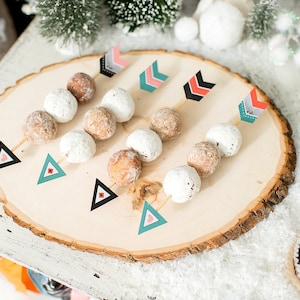 May include: A wooden round platter with a bark edge, decorated with white and brown cake pops on sticks. Each cake pop is topped with a colorful arrow-shaped paper flag.