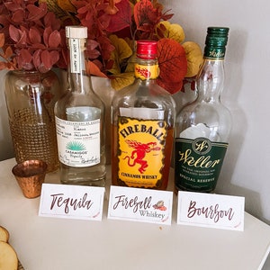 May include: Three bottles of liquor with labels: Tequila, Fireball Cinnamon Whiskey, and Weller Special Reserve Bourbon. The bottles are arranged on a white surface with small signs in front of them.