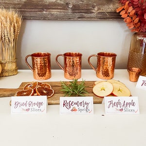 May include: Three copper mugs with handles, a wooden board with dried orange slices, rosemary sprigs, and fresh apple slices.  The board has signs that read "Dried Orange Slices", "Rosemary Sprigs", and "Fresh Apple Slices".  A bottle of tequila and a shot glass are on the counter.