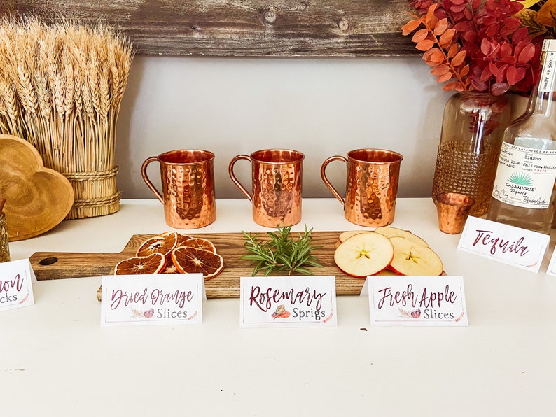May include: Three copper mugs with handles, a wooden board with dried orange slices, rosemary sprigs, and fresh apple slices.  The board has signs that read "Dried Orange Slices", "Rosemary Sprigs", and "Fresh Apple Slices".  A bottle of tequila and a shot glass are on the counter.