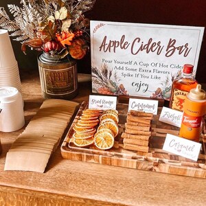 May include: A wooden tray with various fall-themed ingredients for an apple cider bar. The ingredients include cinnamon sticks, dried orange slices, caramel syrup, and Fireball whiskey. A sign reads "Apple Cider Bar" with instructions to "Pour yourself a cup of hot cider, add some extra flavors, spike if you like, enjoy".