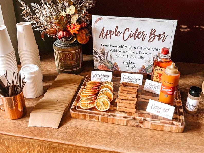 May include: A wooden tray with various fall-themed ingredients for an apple cider bar. The ingredients include cinnamon sticks, dried orange slices, caramel syrup, and Fireball whiskey. A sign reads "Apple Cider Bar" with instructions to "Pour yourself a cup of hot cider, add some extra flavors, spike if you like, enjoy".