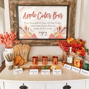 May include: A rustic wooden sign with the text "Apple Cider Bar" and instructions for making a drink. The sign is displayed above a white dresser with copper mugs, various ingredients, and bottles of alcohol. The ingredients include cinnamon sticks, dried orange slices, rosemary sprigs, fresh apple slices, tequila, Fireball whiskey, and bourbon.