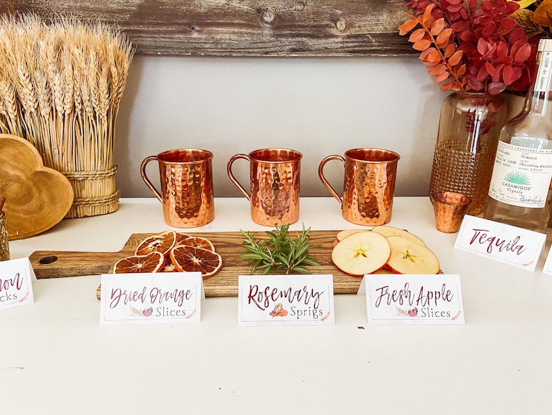 May include: Three copper mugs with handles, a wooden board with dried orange slices, rosemary sprigs, and fresh apple slices.  The board has signs that read "Dried Orange Slices", "Rosemary Sprigs", and "Fresh Apple Slices".  A bottle of tequila and a shot glass are on the counter.