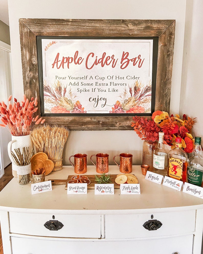 "Apple Cider Bar" Sign with instructions to "Pour yourself a cup of hot cider, add some extra flavors, spike if you like, enjoy". The sign is decorated with fall-themed imagery. A white table with a variety of fall-themed ingredients and drinks for a cider bar, including cinnamon sticks, dried orange slices, rosemary sprigs, fresh apple slices, tequila, fireball whiskey, and bourbon. Printable Sign and Coordinating Labels