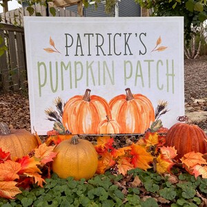 Puede incluir: Un cartel blanco con texto verde que dice "Patrick's Pumpkin Patch". El cartel está rodeado de follaje de otoño y calabazas.