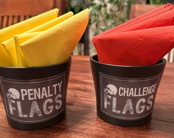 May include: Two black containers with white labels. The container on the left has yellow paper napkins inside and the label says "Penalty Flags". The container on the right has red paper napkins inside and the label says "Challenge Flags".